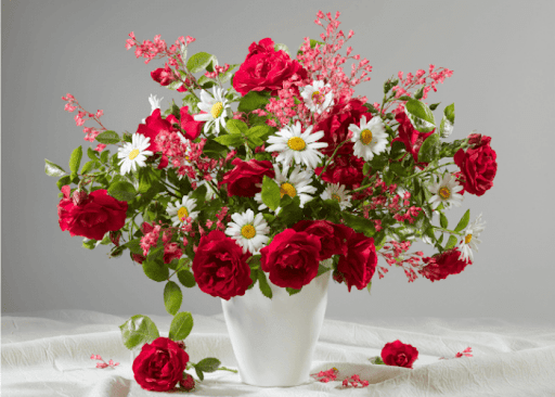A white vase filled with a vibrant floral arrangement of red roses, white daisies, and pink filler flowers, set against a neutral gray background on a white fabric surface.