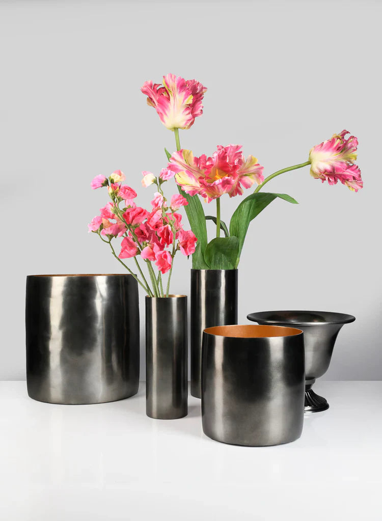 Three gold vases sit on a wooden surface; two hold white flowers with green leaves, while the third vase is empty. The background is a beige textured wall.
